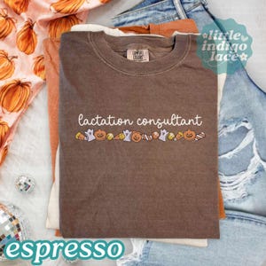 May include: A brown crewneck t-shirt featuring the white text "lactation consultant" above a horizontal row of small, colorful Halloween-themed graphics including ghosts, jack-o'-lanterns, and candy corn. The shirt has a Comfort Colors brand label.