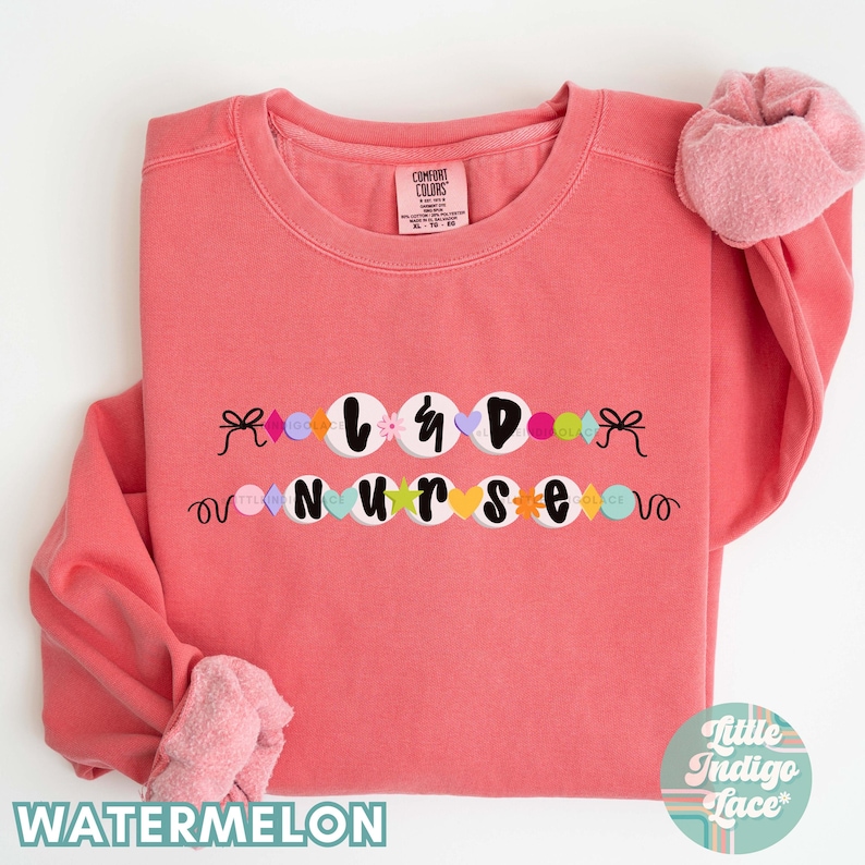 K&ouml;nnte beinhalten: Ein rosa Sweatshirt mit den Worten "L&D Nurse", die in bunten Buchstaben geschrieben sind und mit einer Schnur aus bunten Perlen und Schleifen verziert sind.