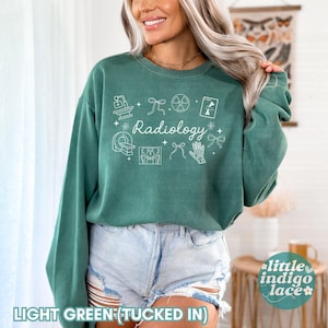 May include: Light green sweatshirt with the word "Radiology" and related medical icons in white. The long-sleeved top has a crew neck and is paired with denim shorts. The text "LIGHT GREEN (TUCKED IN)" is at the bottom.