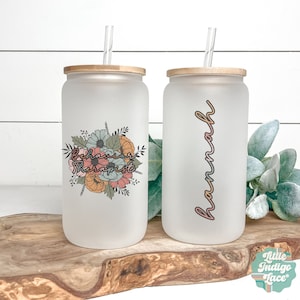 May include: Two frosted glass beverage cups with bamboo lids and clear straws. One cup features a floral design with the words "Behavior Therapist." The other cup has the name "Hannah" in a colorful script. Both cups are on a wooden surface.