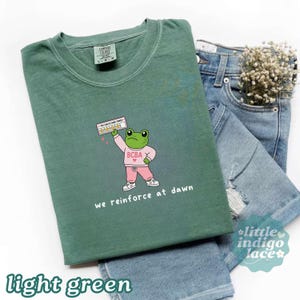 May include: A sage green crewneck t-shirt featuring a graphic of a green frog wearing a pink sweatshirt with the text BCBA and pink sweatpants. The frog holds a sign that says YOU CAN DO HARD THINGS with five stars. The text reads we reinforce at dawn.