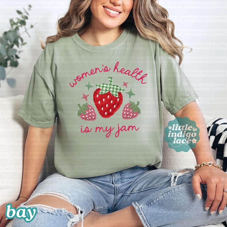 May include: A sage green short-sleeve t-shirt featuring a graphic of three strawberries with a green gingham pattern on the leaves. The text reads "women's health is my jam" in a red script font, accompanied by small red and green star accents.