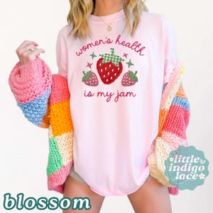 May include: A light pink t-shirt featuring a graphic of three strawberries with green and red checkered patterns. The text reads "women's health is my jam" in red cursive lettering. The shirt is worn under a chunky, multi-colored knit cardigan.