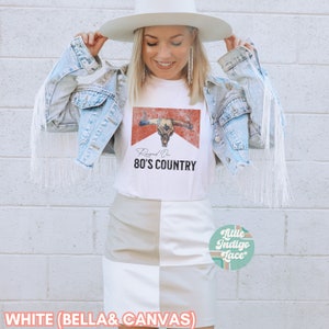 May include: White t-shirt with a distressed red and orange graphic of a bull skull with the text "Raised On 80's Country". The model is wearing a denim jacket with white fringe and a white and beige skirt.