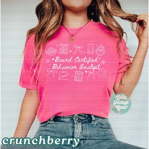 May include: A bright pink t-shirt with the words "Board Certified Behavior Analyst" in white, along with related icons. The word "crunchberry" is at the bottom. The shirt is a casual style.