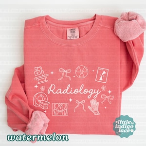 May include: A watermelon-colored sweatshirt with the word "Radiology" in a script font, along with white line art illustrations of medical imaging equipment and symbols. The sweatshirt has a Comfort Colors tag.