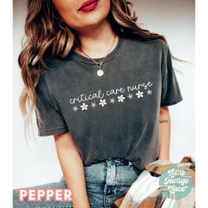 Puede incluir: Una camiseta gris oscuro con el texto "critical care nurse" en escritura blanca con peque&ntilde;as flores blancas con centros amarillos. La camiseta la lleva puesta una persona con el pelo largo y casta&ntilde;o.