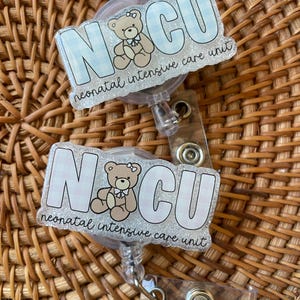 May include: Two badge reels with the letters "NCU" in a light blue gingham pattern, featuring a teddy bear illustration. The text "neonatal intensive care unit" is below the letters. The reels have a clear plastic retractable cord and a metal clip.