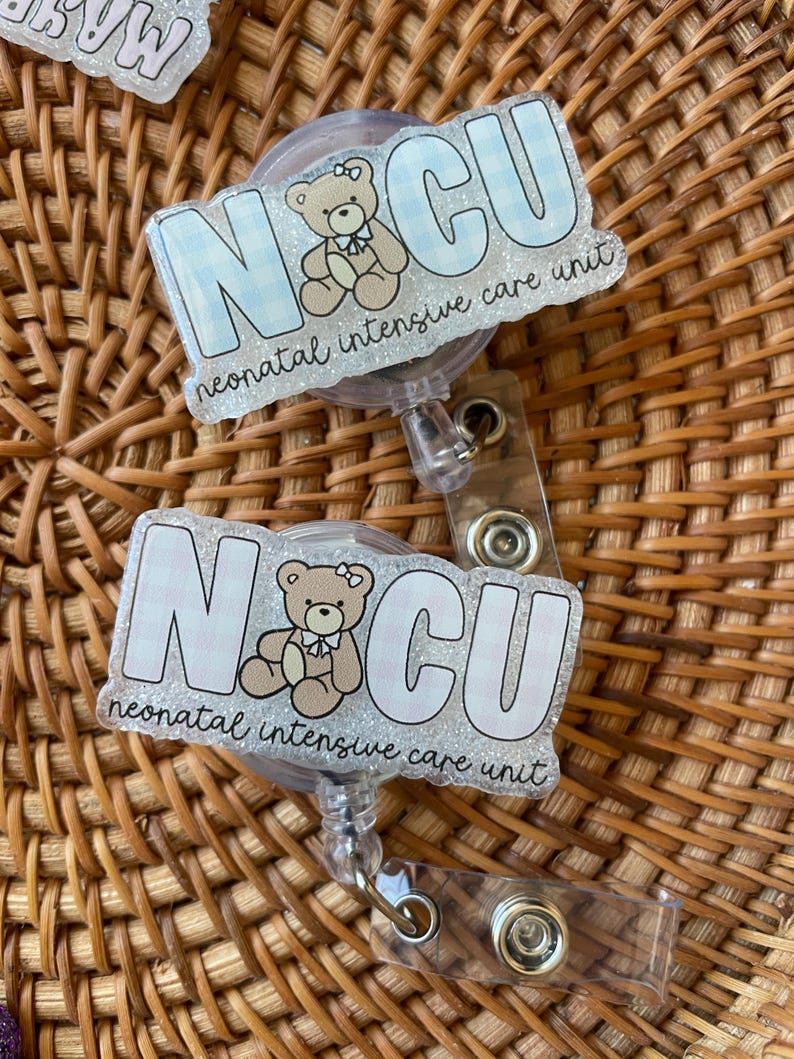 May include: Two badge reels with the letters "NCU" in a light blue gingham pattern, featuring a teddy bear illustration. The text "neonatal intensive care unit" is below the letters. The reels have a clear plastic retractable cord and a metal clip.