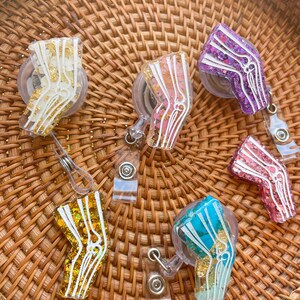 Pu&ograve; includere: Sei porta badge retrattili di diversi colori con una custodia in plastica trasparente e un design di osso del ginocchio anatomico bianco. Ogni porta badge ha un glitter di colore diverso incorporato nella plastica. I colori sono oro, rosa, viola, turchese, blu e arancione.
