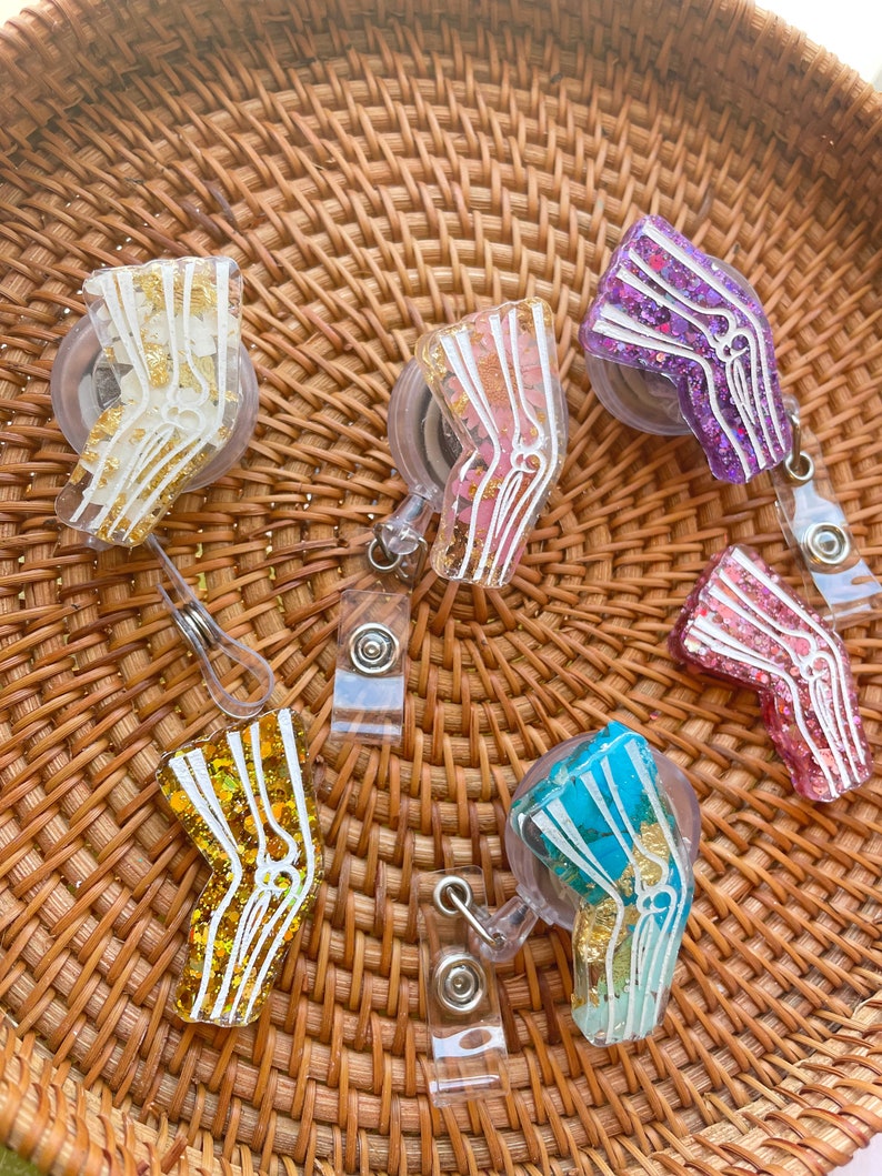 Pu&ograve; includere: Sei porta badge retrattili di diversi colori con una custodia in plastica trasparente e un design di osso del ginocchio anatomico bianco. Ogni porta badge ha un glitter di colore diverso incorporato nella plastica. I colori sono oro, rosa, viola, turchese, blu e arancione.