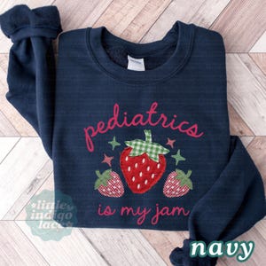 May include: A navy blue crewneck sweatshirt featuring the red text "pediatrics is my jam" and an appliqu&eacute; design of three strawberries with green and white checkered leaves. The strawberries are red with white dots and checkered patterns.