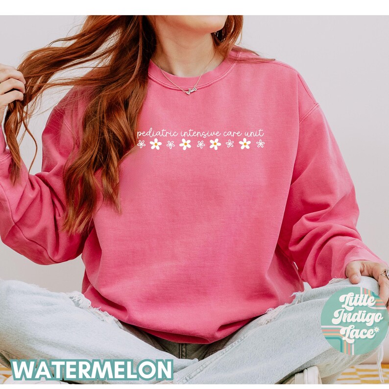 K&ouml;nnte beinhalten: Ein rosa Sweatshirt mit dem Text "pediatric intensive care unit" und einer Reihe wei&szlig;er Blumen, die auf der Vorderseite aufgedruckt sind.