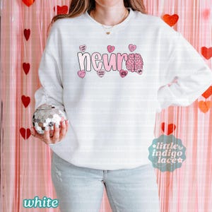 May include: White sweatshirt with the word "neuro" in a pink gingham font, a pink brain graphic, and pink heart-shaped candies with phrases like "I love you" and "neuro crew." The sweatshirt has long sleeves.