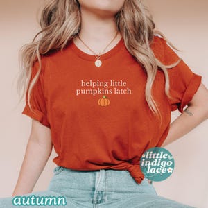 May include: An orange t-shirt with the text "helping little pumpkins latch" and a small pumpkin graphic. The shirt is a warm, autumnal color and is paired with blue jeans. The words "autumn" and "little indigo lace" are also visible.