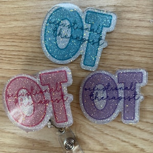 May include: Three glittery acrylic badge reels shaped like the letters OT. One is blue, one is pink, and one is purple. Each piece features the text occupational therapist printed in a dark cursive font across the center of the letters.
