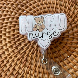 May include: A white and pink glitter badge with the text "NICU nurse" and a small brown teddy bear with a pink bow. The badge has a clear plastic retractable badge holder with a silver clip.
