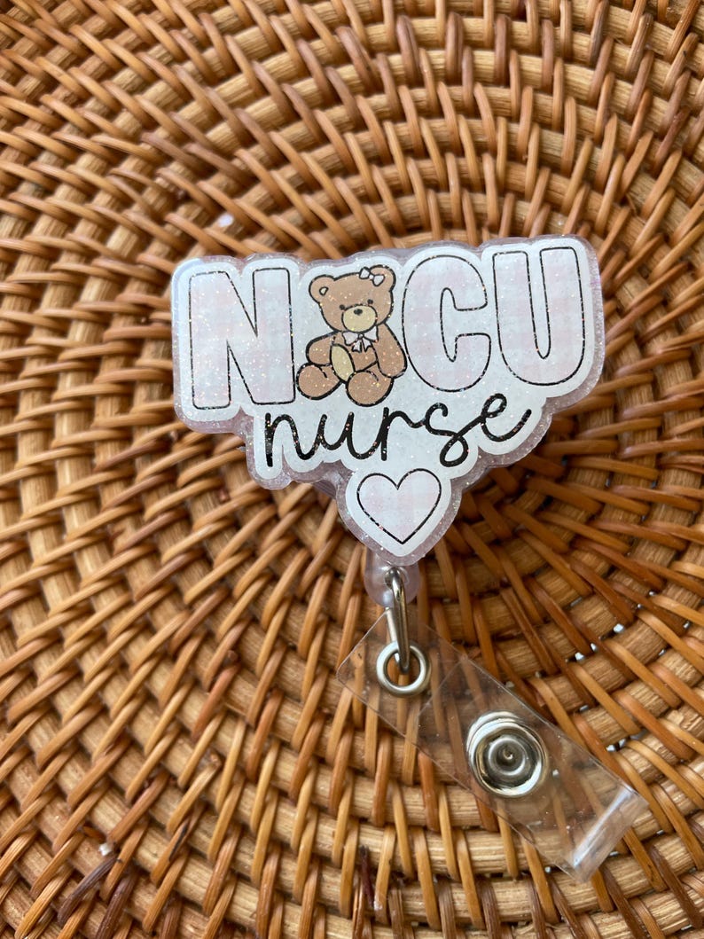 May include: A white and pink glitter badge with the text "NICU nurse" and a small brown teddy bear with a pink bow. The badge has a clear plastic retractable badge holder with a silver clip.