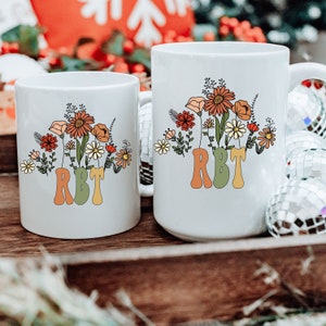 May include: Two white ceramic mugs with a floral design and the text "RBT" in a retro font. The flowers are in shades of orange, yellow, and green.