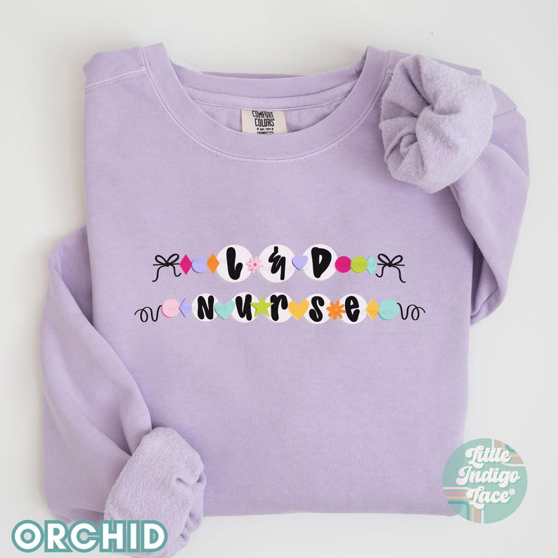 K&ouml;nnte beinhalten: Ein lavendelfarbener Pullover mit den Worten "L&D Nurse", die in bunten Kreisen und einem Bogen an jedem Ende ausgeschrieben sind.