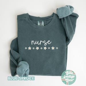 May include: A teal blue sweatshirt with the word "nurse" in white script and a row of white daisies with yellow centers. The sweatshirt is made by "Comfy Colors" and has a "Little Indigo Lace" tag.