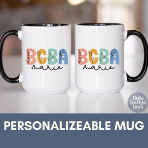 May include: Two white ceramic mugs with black handles. The mugs have a black rim and feature the text "BCBA marie" in colorful letters with floral accents.