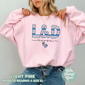 May include: Light pink sweatshirt with the text "L&D nurse" in blue and red stripes with a pair of baby footprints below the text.
