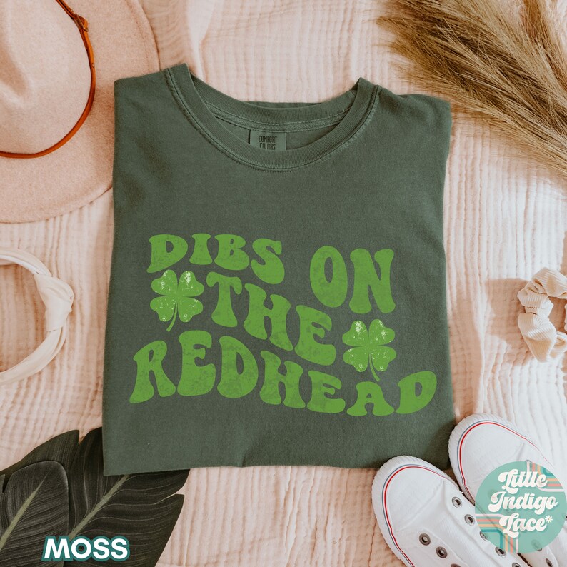Op de afbeelding: Een groen T-shirt met de tekst "DIBS ON THE REDHEAD" in een retro-lettertype met klavertjevier-accenten.