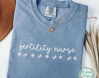 Puede incluir: Una camiseta azul vaquero con el texto "fertility nurse" en letras blancas. Hay pequeñas huellas de pies rosas y azules y flores blancas impresas debajo del texto. La camiseta está hecha por Comfort Colors y tiene una etiqueta que dice "S - P - CH".
