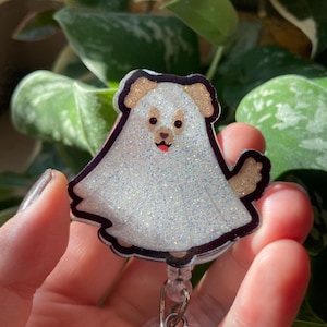 May include: A badge reel featuring a glittery white ghost costume worn by a tan dog. The dog has brown ears, a black nose, and a small red tongue visible. The ghost costume is outlined in black, and the entire piece has a sparkling finish.
