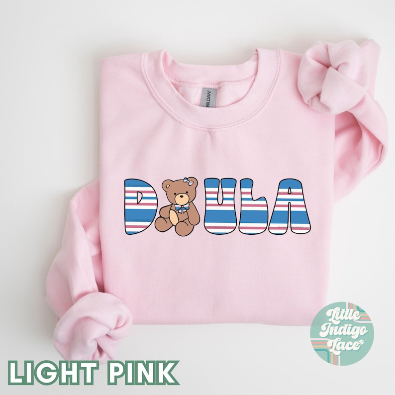 May include: Light pink sweatshirt with the word "DOULA" in blue, red, and white stripes. A small brown teddy bear with a blue bow is in the middle of the word.