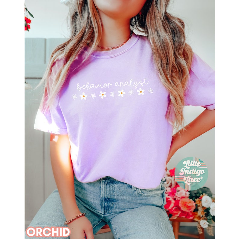 May include: A purple t-shirt with the text "behavior analyst" and a row of white daisies printed on the front. The shirt is being worn by a person with long blonde hair. The text "Little Indigo Face" is visible in the lower right corner of the image.