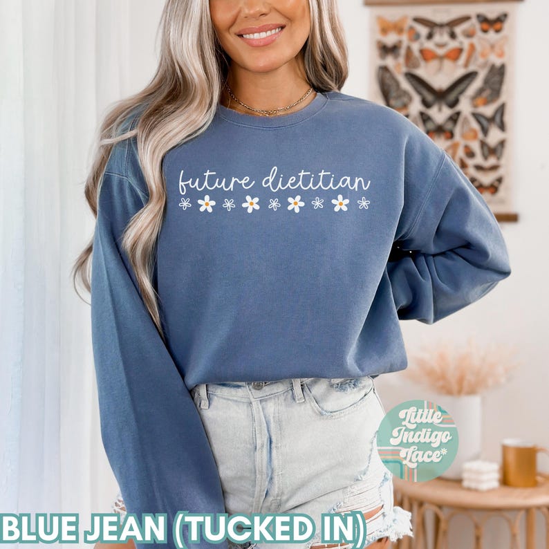 May include: A blue jean colored sweatshirt with the text "future dietitian" in white script, decorated with small white daisy graphics. The sweatshirt is a long-sleeved crew neck style.