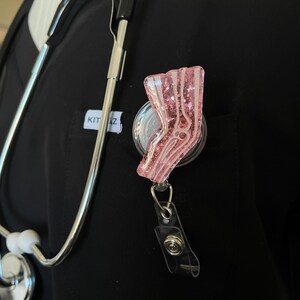 Pu&ograve; includere: Portabadge con paillettes rosa con un designo di osso di gamba anatomico bianco. Il portabadge &egrave; attaccato a un laccio nero con un fermaglio argentato.