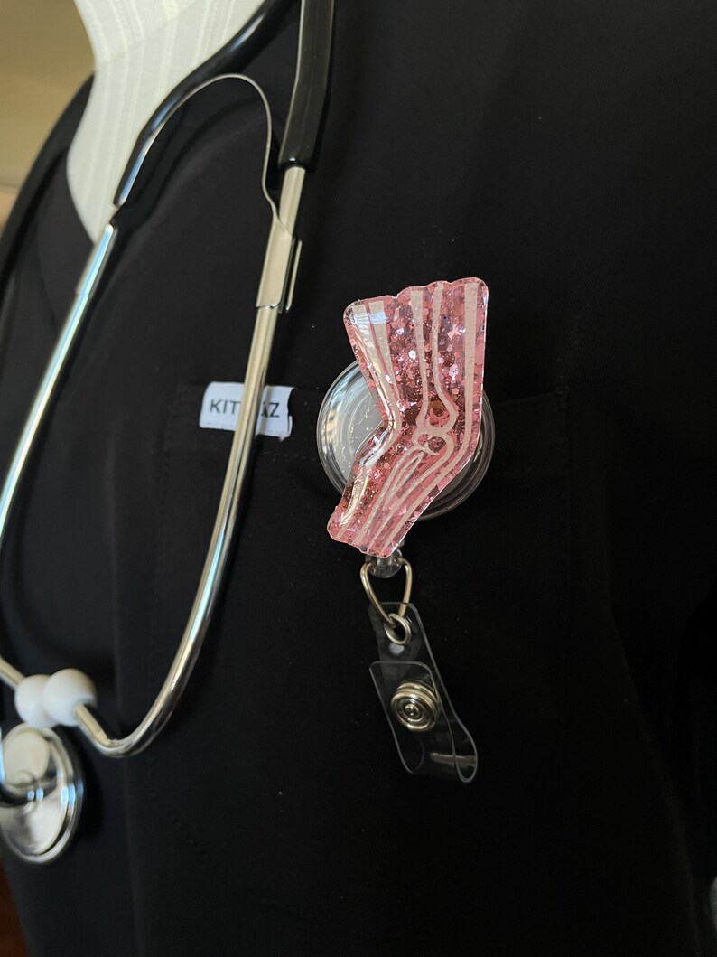 Pu&ograve; includere: Portabadge con paillettes rosa con un designo di osso di gamba anatomico bianco. Il portabadge &egrave; attaccato a un laccio nero con un fermaglio argentato.