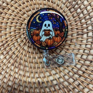 May include: A circular badge reel featuring a stained-glass style illustration of a white ghost holding a jack-o'-lantern mug. The scene includes orange pumpkins, autumn leaves, a crescent moon, and a dark blue starry sky with glitter accents.