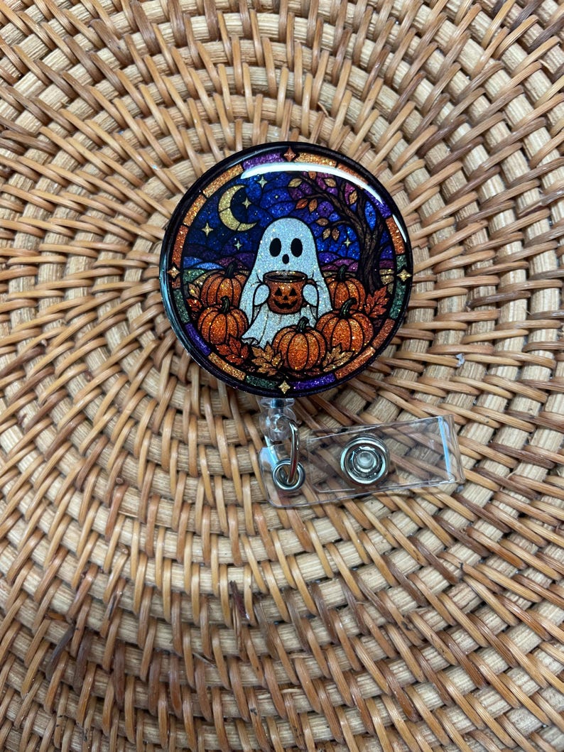 May include: A circular badge reel featuring a stained-glass style illustration of a white ghost holding a jack-o'-lantern mug. The scene includes orange pumpkins, autumn leaves, a crescent moon, and a dark blue starry sky with glitter accents.