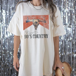 May include: A white oversized t-shirt with a distressed red and orange graphic of a bull skull with the text "Raised On 90's Country" printed on the front.