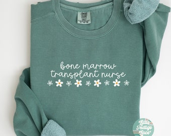 May include: Light green sweatshirt with the text "bone marrow transplant nurse" in white lettering and a row of white daisies with yellow centers.