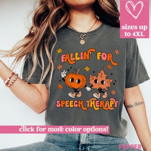 May include: Dark gray t-shirt with the text "FALLIN' FOR SPEECH THERAPY" in orange and pink. The shirt features a cartoon pumpkin and leaf holding hands. The text "sizes up to 4XL" is in a pink heart-shaped box.