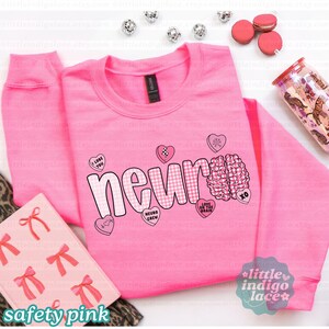 May include: A safety pink sweatshirt with the word "neur" in a checkered pattern, a brain graphic, and heart-shaped candies. The sweatshirt also features the phrases "I love you", "Neuro Crew", and "Love on the brain".