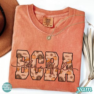 May include: A coral-colored t-shirt with the word "BOBA" in large, block letters filled with a Thanksgiving-themed pattern of turkeys, pumpkins, and pie slices. The word "thankful" is written in cursive below.