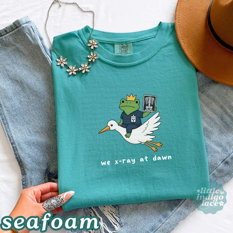 May include: A teal short-sleeved t-shirt featuring a graphic of a frog wearing a crown and scrubs, holding an X-ray while riding a white stork. The text "we x-ray at dawn" is printed below the graphic. The shirt has a crew neckline and a tag.