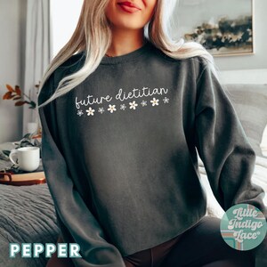 May include: A dark gray sweatshirt with the words "future dietitian" in white script, decorated with small white and yellow flower accents. The word "PEPPER" is printed in white at the bottom left corner.