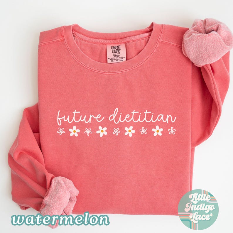 May include: A coral-colored sweatshirt with the words "future dietitian" in white script, decorated with small white daisy graphics. The word "watermelon" is printed in a teal font at the bottom. A pink scrunchie is on the sleeve.