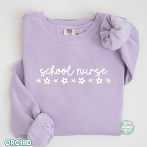 May include: A lavender sweatshirt with the text "school nurse" in white lettering and a row of white daisies below. The sweatshirt has a crew neck and long sleeves.
