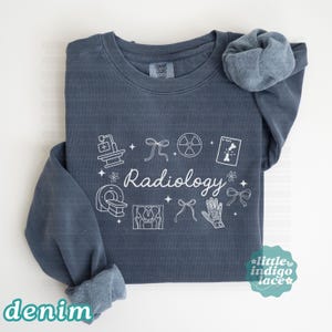 May include: Denim blue sweatshirt with white "Radiology" text and related medical illustrations. Features a crew neck and long sleeves, with the word "denim" at the bottom left. The sweatshirt has a relaxed fit and a soft, comfortable appearance.
