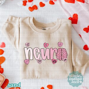 May include: Sand-colored sweatshirt with the word "neuro" in pink gingham letters. Pink heart-shaped candies with phrases like "I love you" and a brain graphic are also featured. The sweatshirt is surrounded by heart-shaped decorations.