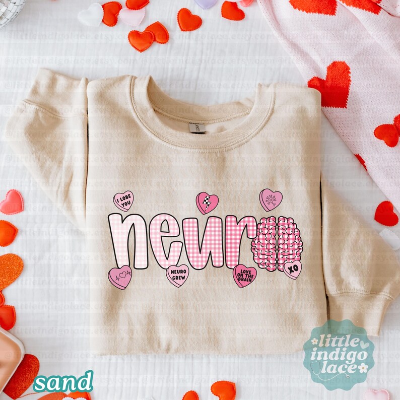 May include: Sand-colored sweatshirt with the word "neuro" in pink gingham letters. Pink heart-shaped candies with phrases like "I love you" and a brain graphic are also featured. The sweatshirt is surrounded by heart-shaped decorations.
