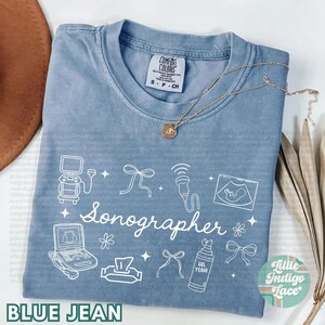 May include: Blue jean colored t-shirt with the word "Sonographer" in white script. The shirt features white line drawings of sonography equipment, bows, and the text "GEL YEAH!". A gold necklace with a pendant is draped across the shirt.